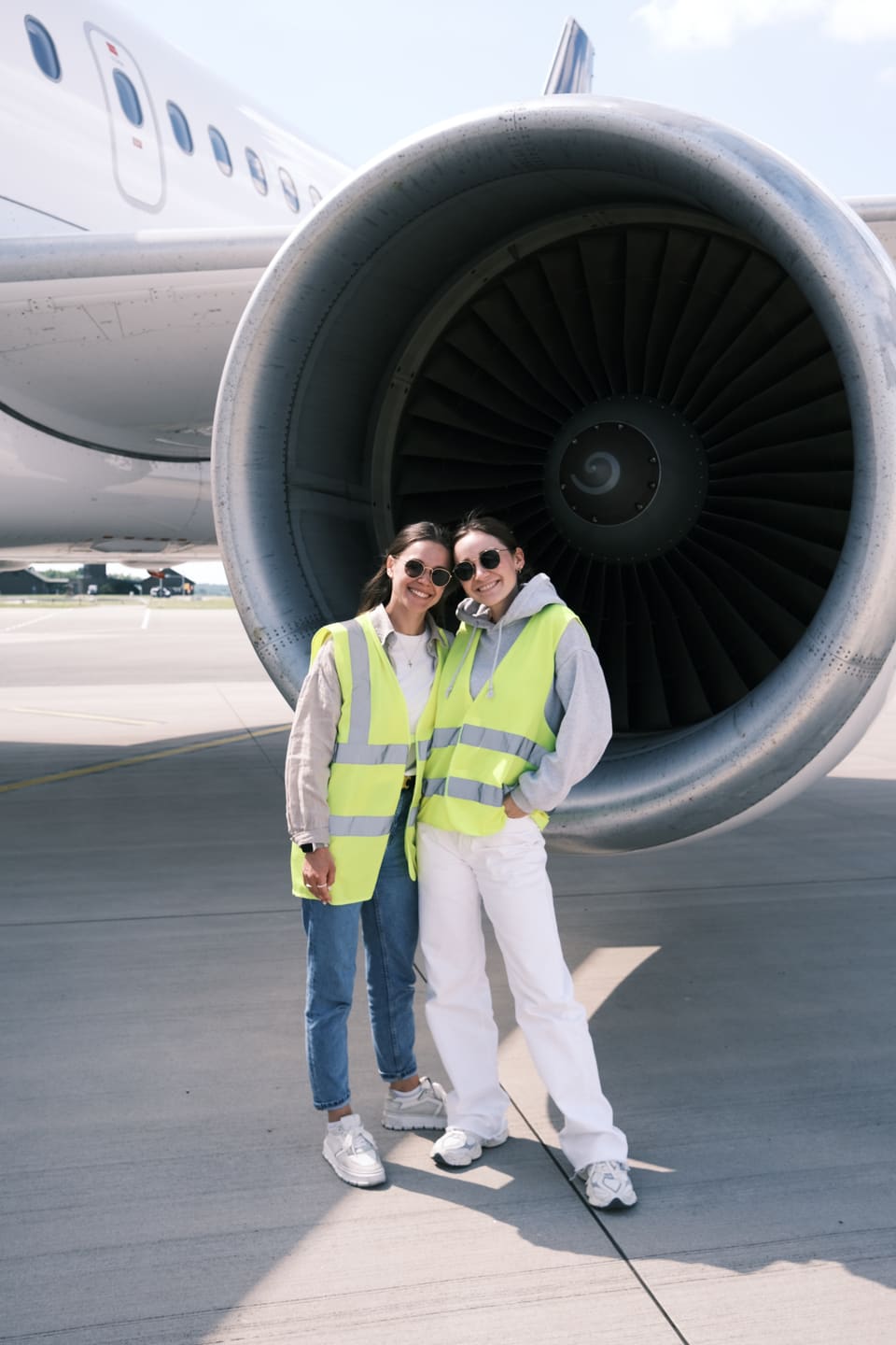 Anna und Julia in Warnwesten und mit Sonnenbrillen auf dem Vorfeld, Arm in Arm vor dem Triebwerk eines Flugzeugs.